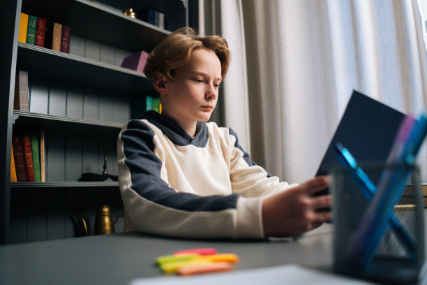特写的焦点严肃的小学生男孩独自坐在书桌上阅读纸质书在光儿童房靠近窗户。图片下载