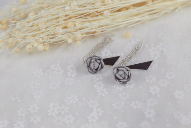 灰色吊坠耳环，它的形状是一朵花。在耳环旁边放一束花的标本。这些东西都放在薄纱上。这对耳环看起来非常优雅时尚。耳环以流苏和金属三角形装饰。图片下载