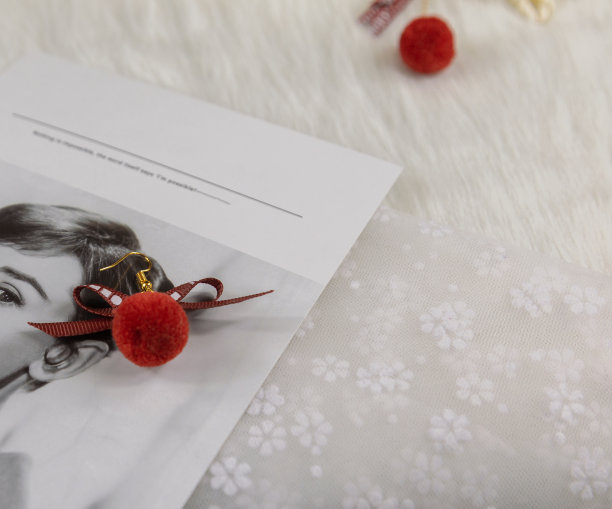 红色的绒球耳环，它的形状是一个毛茸茸的球。耳环旁边有装饰物品，如纸板和花束标本。这些东西都放在薄纱上。耳环看起来非常优雅和时尚。图片下载