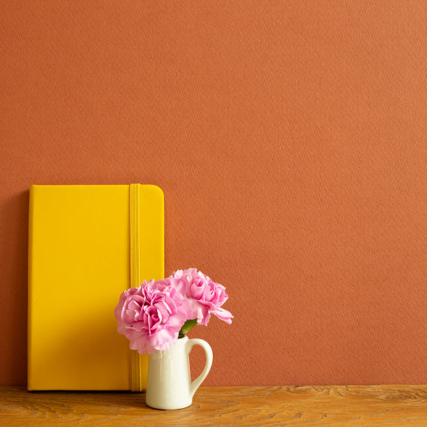 黄色笔记本和粉红色康乃馨花放在木桌上。棕色的背景图片下载