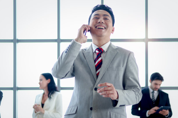 资深亚洲微笑的商人老板西装打电话，笑在窗口办公室工作与商务人员的同事作为模糊的背景。图片下载