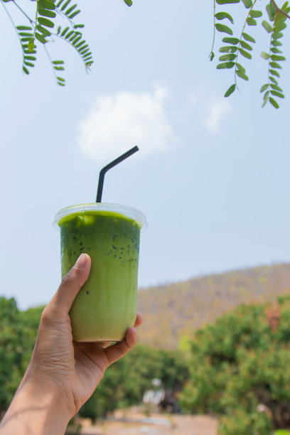 一个女人的手拿着一个塑料杯，自制抹茶绿茶。它的背景是群山和树木。图片下载