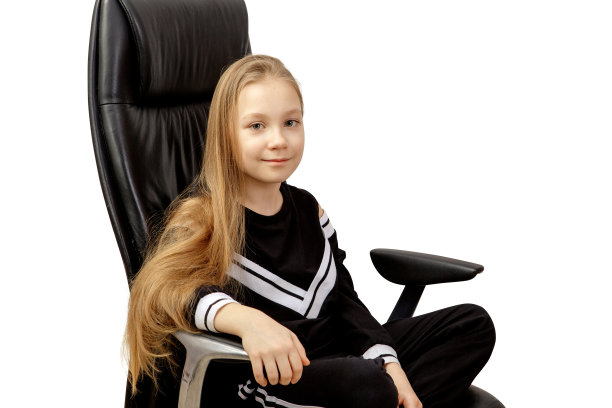 红头发的漂亮微笑的小女孩坐在黑色的办公椅上。孤立在白色背景上。剪切路径。图片下载