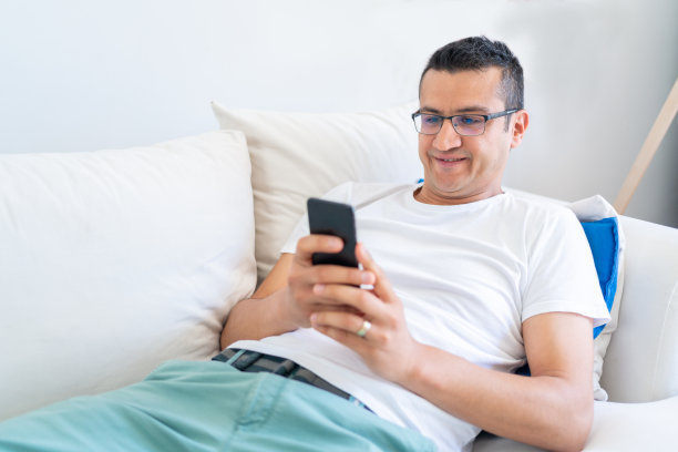 躺在沙发上用智能手机的快乐微笑的男人。在家放松图片下载