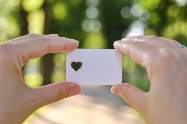 在阳光下，一个女孩的手里拿着一张白色的心形卡片。特写的明信片。形式的祝贺。浪漫的邀请。模型。为信息或邀请腾出空间图片下载