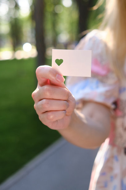在阳光下，一个女孩的手里拿着一张白色的心形卡片。特写的明信片。形式的祝贺。浪漫的邀请。模型。为信息或邀请腾出空间图片下载