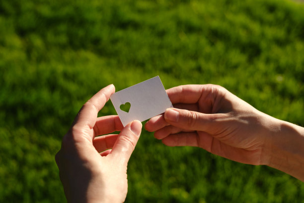 在阳光下，一个女孩的手里拿着一张白色的心形卡片。特写的明信片。形式的祝贺。浪漫的邀请。模型。为信息或邀请腾出空间图片下载