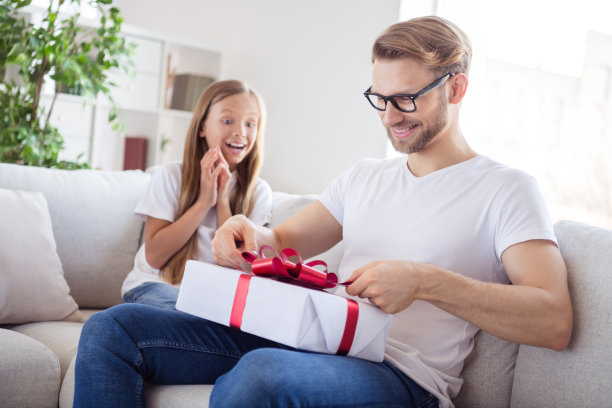 有趣的照片兴奋的父亲女儿穿着休闲衣服坐在沙发拆礼物盒室内房子公寓图片下载