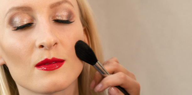 一个美丽的年轻金发女人的特写与闭上眼睛的晚妆与红色的口红在她的嘴唇与化妆师的手与粉刷复制空间。注意嘴唇图片下载