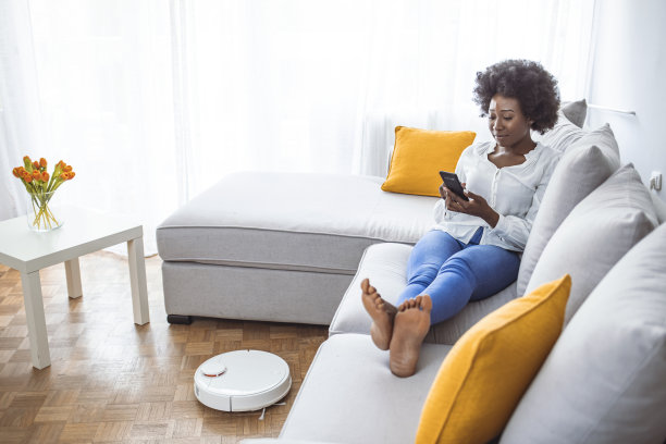 当女人在沙发上休息时，机器人真空吸尘器正在打扫房间。图片下载