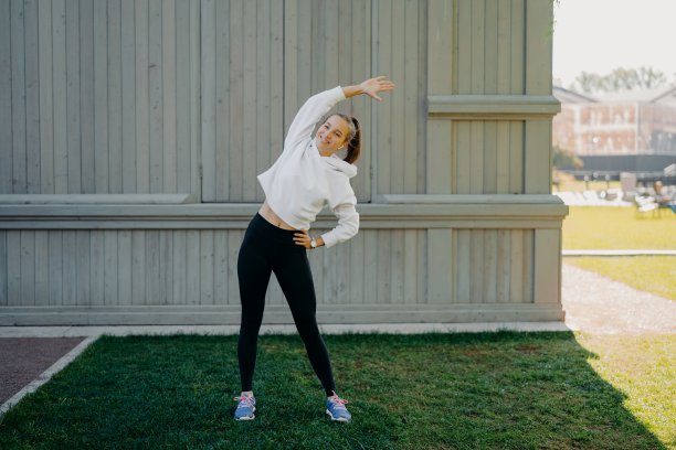 全长镜头的活跃的女运动员在户外锻炼，画身体斜靠在不同的一边，穿着运动衫长裤和运动鞋，在耳机里听音乐，有冷冷的身体愉快地微笑图片下载