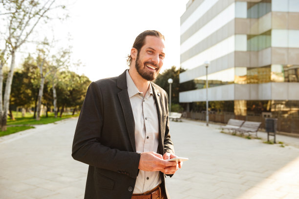 照片中的白人微笑的男人30多岁的商务西装看着你和拿着智能手机，而走在办公大楼附近图片下载