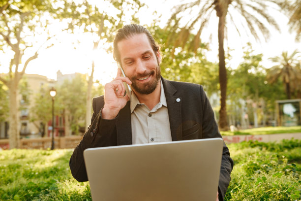 照片的特写，像商业英俊男子与扎着头发工作银色笔记本和有移动电话，而坐在城市公园在阳光明媚的一天图片下载