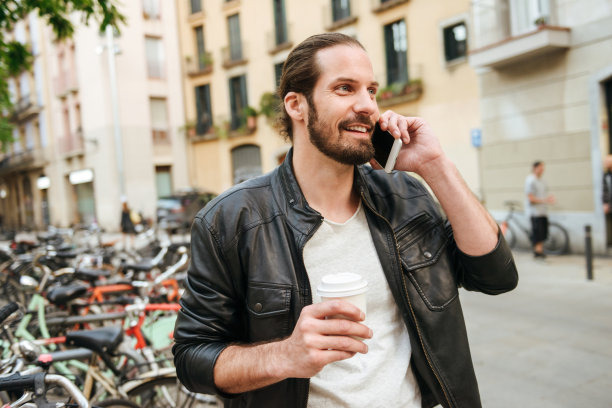 照片中，30多岁的帅哥穿着休闲，拿着外卖咖啡，在城市街道上用手机微笑着交谈图片下载