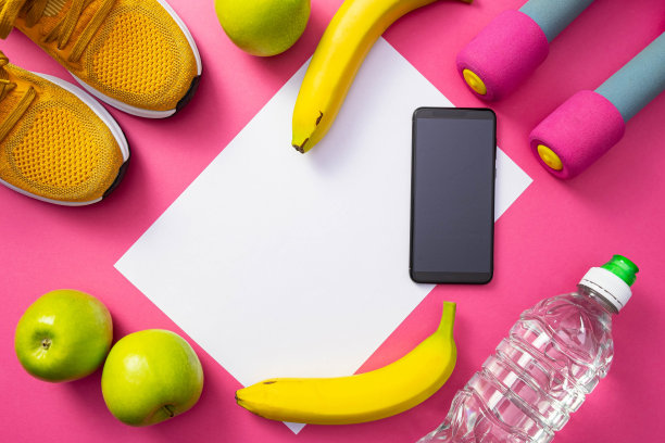 一个手机、一瓶水、哑铃、黄色运动鞋、苹果和香蕉放在普通的粉色背景上图片下载