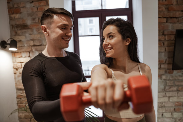 年轻的白人男子用哑铃向年轻的黑发女子展示锻炼。这对夫妇在家喜欢运动。在客厅的家庭氛围中锻炼图片下载