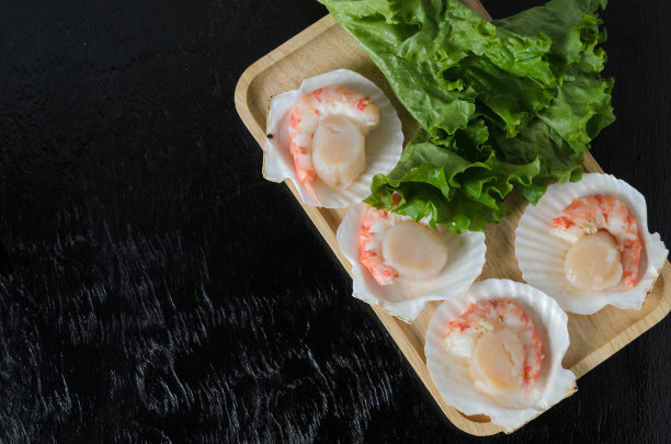 一个天然的生扇贝，贝壳里有虾尾，木碗里有一片莴苣叶，背景是黑色的图片下载