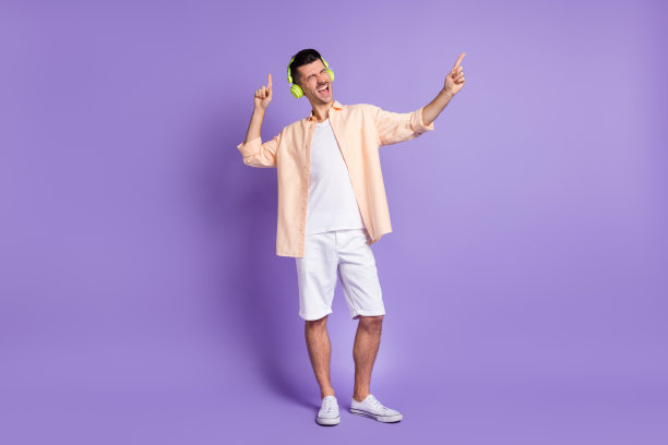 照片上的家伙闭上眼睛听音乐指指点点唱戴耳机米色衬衫孤立的紫罗兰色背景图片下载