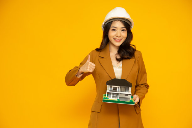亚洲女性或工程师竖起大拇指，戴着白色头盔，拿着独栋住宅或独立住宅模型，在黄色背景上孤立图片下载