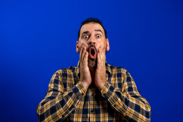 胡子男子在黄色格子衬衫与手在他的脸，嘴和大眼睛在恐惧姿态孤立在蓝色工作室背景图片下载