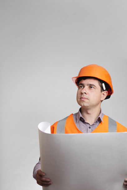 在灰色的工作室背景上穿着制服的人与开放的纸质建筑计划。建筑工人戴着安全帽，耳朵后面有记号笔图片下载