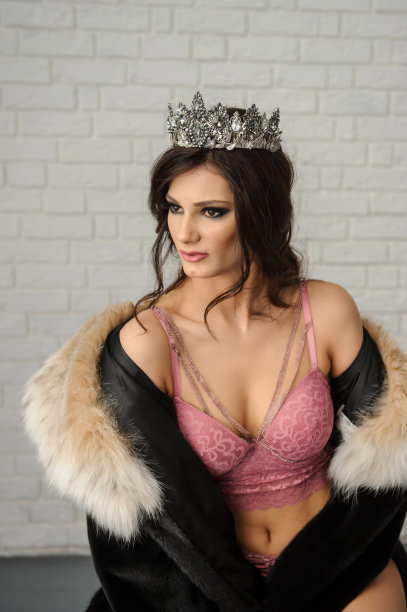 照片中的一个性感的时尚女人在黑色皮草大衣米色领子，在粉红色内衣与皇冠上她的头坐在一个椅子上的白色砖背景图片下载