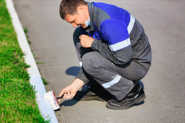 一个夏天，一个穿着工作服的工人在用白色油漆粉刷路边。城市服务，改善领土图片下载