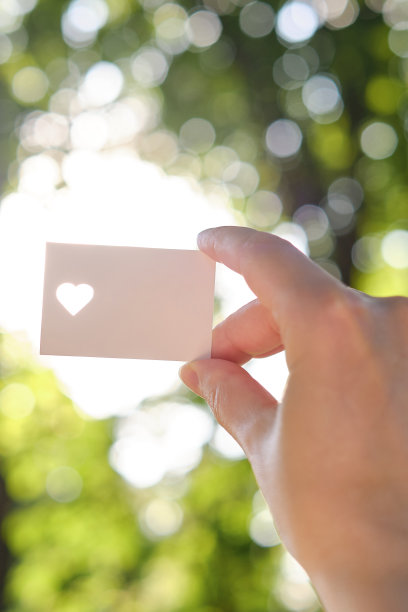 在阳光下，一个女孩的手里拿着一张白色的心形卡片。特写的明信片。形式的祝贺。浪漫的邀请。模型。为信息或邀请腾出空间图片下载