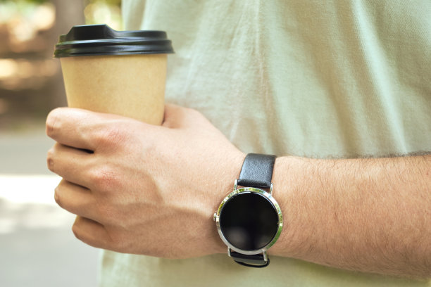 手握咖啡纸杯的人手里拿着一个时钟。一次性纸杯靠近。一个男人看他时髦的手表上的时间。时间管理，机动性，社会和技术概念图片下载