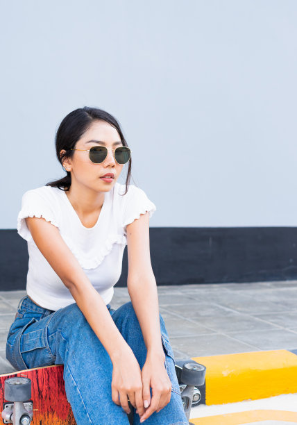 亚洲可爱的女孩或女人坐在街上，把她的腿放在滑板上，在日光下，暑假，在有复印空间的酷墙前看起来空空如也图片下载