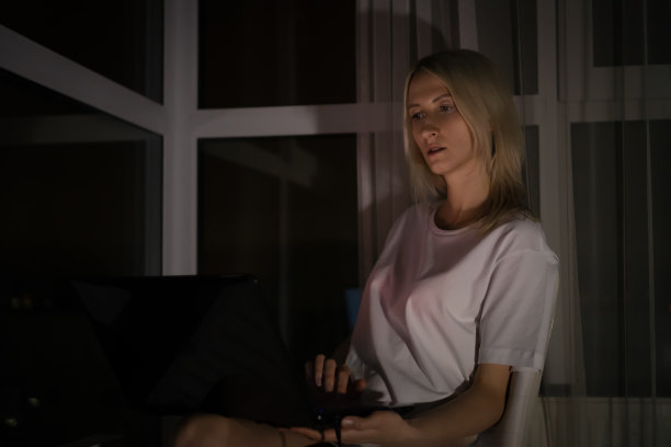 年轻的金发女郎穿着白衬衫，晚上在窗边用笔记本电脑工作图片下载