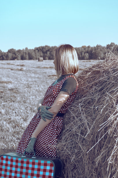 穿着豌豆裙的女孩斜着身子，手里拿着一个方格手提箱，倚靠在一卷干草上。农业的地形。复古的风格图片下载