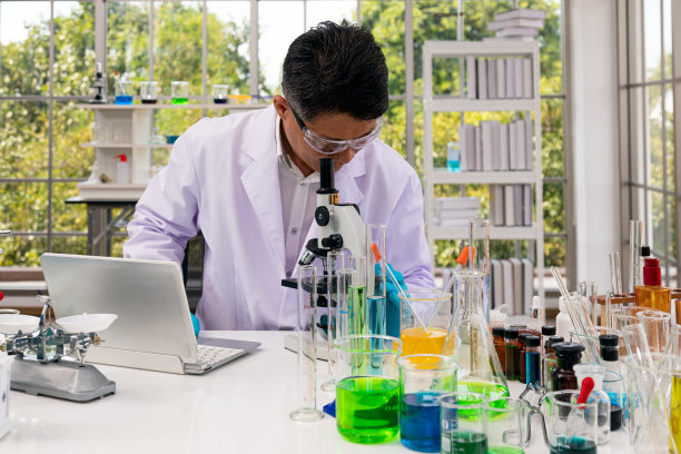 资深科学家在实验室工作期间，戴着安全眼镜观察显微镜，检查笔记本电脑上的实验数据图片下载