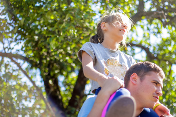 散了焦的哥哥骑在姐姐的背上。快乐女孩的肖像在男人的肩膀上，背着。女孩飞。家人在户外玩耍。绿树模糊的背景。特写镜头。侧视图。的焦点图片下载