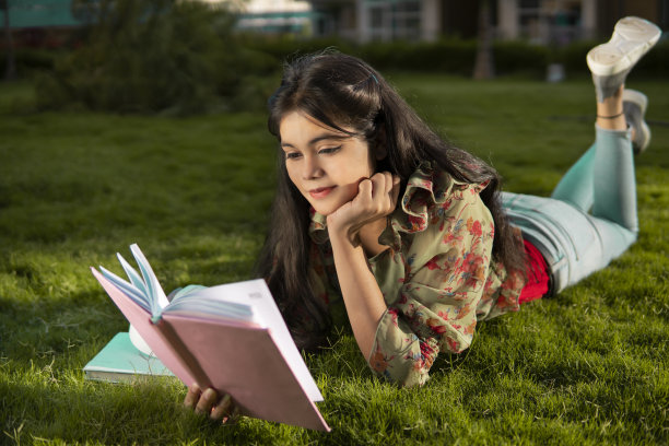 美丽的年轻女子躺在公园的草地上，在白天的新鲜空气中看书图片下载