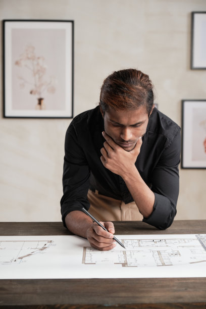 专注于年轻的印度建筑设计师在黑衬衫倚在木桌上，手握下巴，而审查房屋计划图片下载