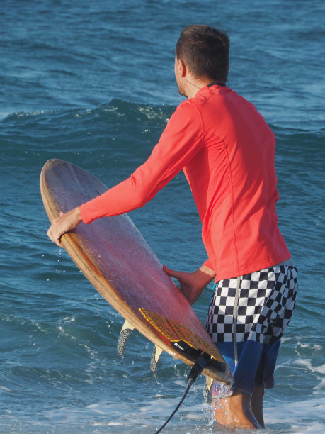 冲浪男子穿着红色长袖短裤，背着冲浪板，进入海水中。积极的生活方式。后视图。图片下载