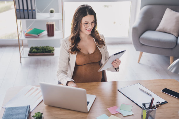 孕妇手持平板电脑在现代办公桌旁用笔记本电脑工作的照片图片下载