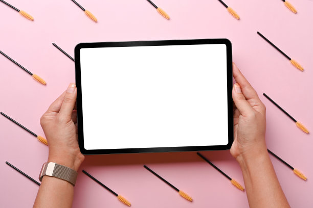 女人拿着数字平板电脑与眉毛和睫毛刷在粉红色的背景。图片下载