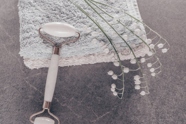 玉石面部罗拉提肤调色工具。白色美丽的脸辊，按摩器和百合的山谷花，毛巾在灰色的背景。图片下载