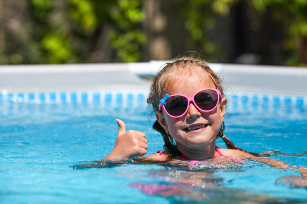 在一个阳光明媚的日子里，可爱的小女孩戴着太阳镜在游泳池里微笑。孩子竖起大拇指，像是在看相机。暑假图片下载