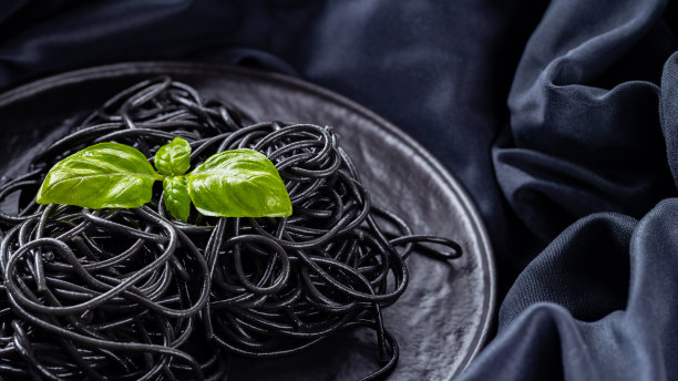黑色的意大利面和罗勒在深色的背景上。黑色意大利面，墨鱼墨，黑布上。黑色盘子上放有新鲜罗勒的意大利面。副本的空间。俯视图图片下载