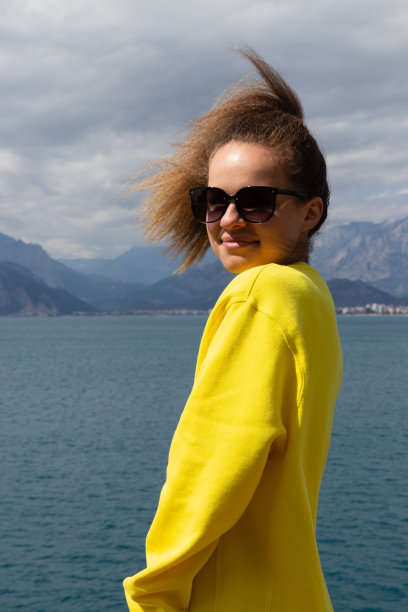 照片上的年轻女孩穿着黄色运动衫，戴着太阳镜，面带微笑，站在海边享受生活。图片下载