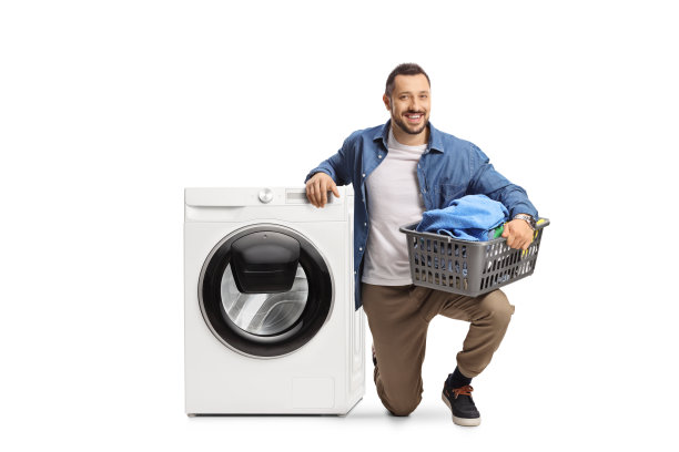 一个微笑的年轻人跪在洗衣机旁边，拿着一个洗衣篮图片下载