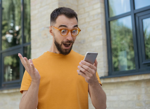 兴奋的男人用手机上网购物，体育博彩，交流，站在户外。年轻的留着胡子的自由作家，读着好消息，拿着报酬图片下载