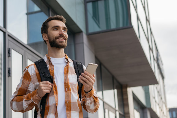 微笑的英俊男子在户外使用手机的肖像。留着胡子的年轻游客背着背包走在城市的大街上，寻找最好的路。旅游的概念图片下载