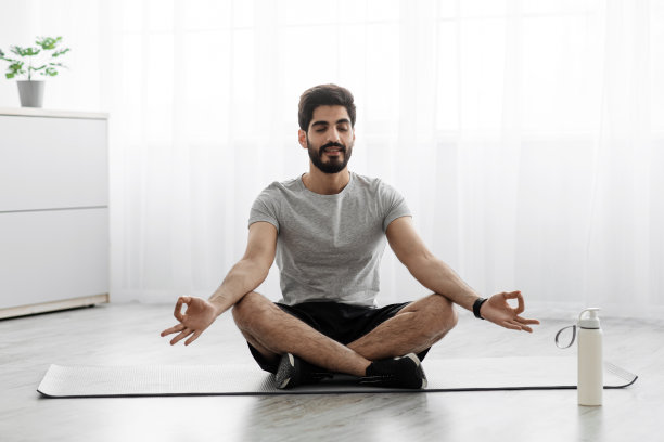 快乐的阿拉伯胡子千禧男子坐在垫子上莲花的位置，休息在地板上与一瓶水图片下载