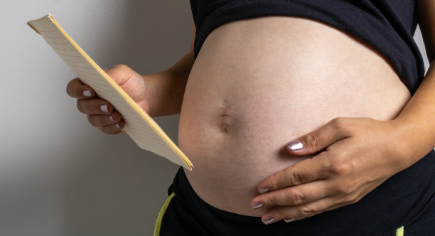一个女人的肚子里怀着一个男孩，手里拿着一张纸，上面列着购买物品的清单和对婴儿未来的货币计算。提到经济和孩子。图片下载