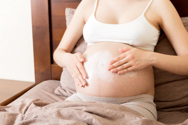 一个怀孕的女孩坐在家里的床上，把抗妊娠纹霜涂在她的肚子上。怀孕、母性、准备和期望的概念图片下载