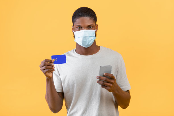简单的支付。非裔美国人戴着面具在黄色背景下使用智能手机和信用卡图片下载